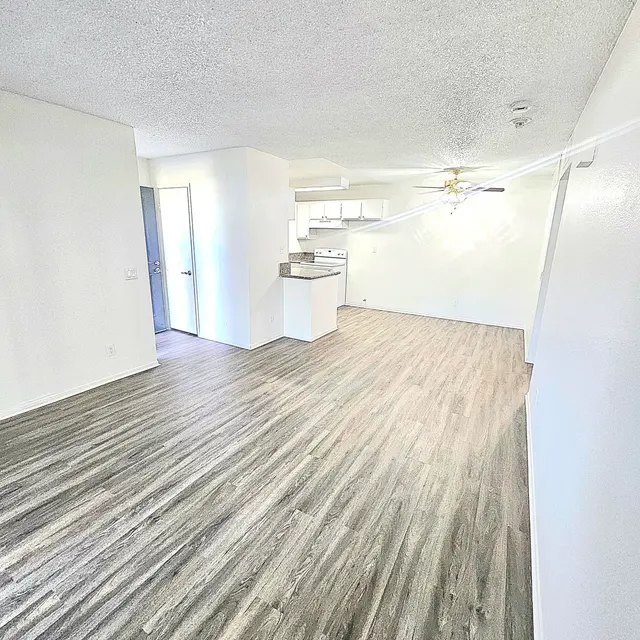 An empty living room with laminate flooring, white walls, and a ceiling fan. A doorway is visible leading to another room, and the kitchen area can be seen in the background.