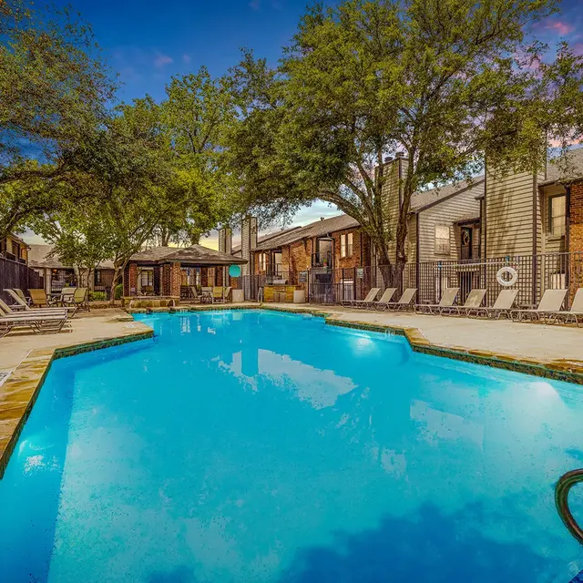 A tranquil apartment complex pool area featuring a clear blue pool surrounded by lounge chairs and trees, under a colorful sky at dusk.
