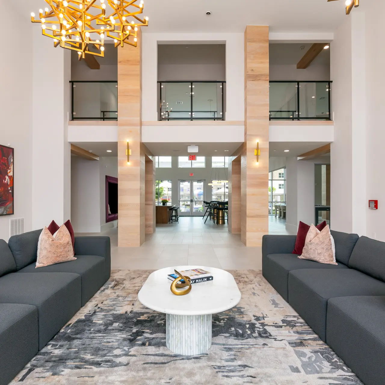 A spacious and modern lounge area featuring two gray sofas with red and white pillows, a round coffee table, and a large window behind. The ceiling is high with stylish light fixtures and wood accents.