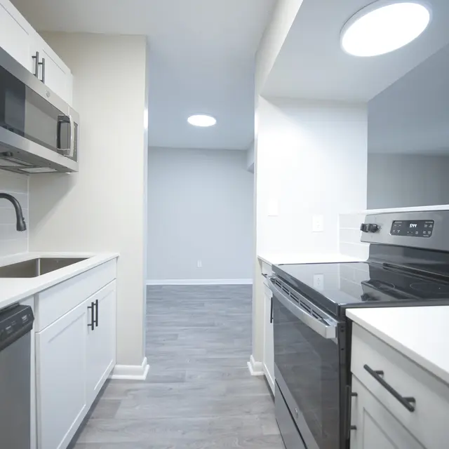 A modern kitchen featuring white cabinetry, stainless steel appliances, and a view of an open living space. The kitchen has a sink, microwave, oven, and a dark countertop.