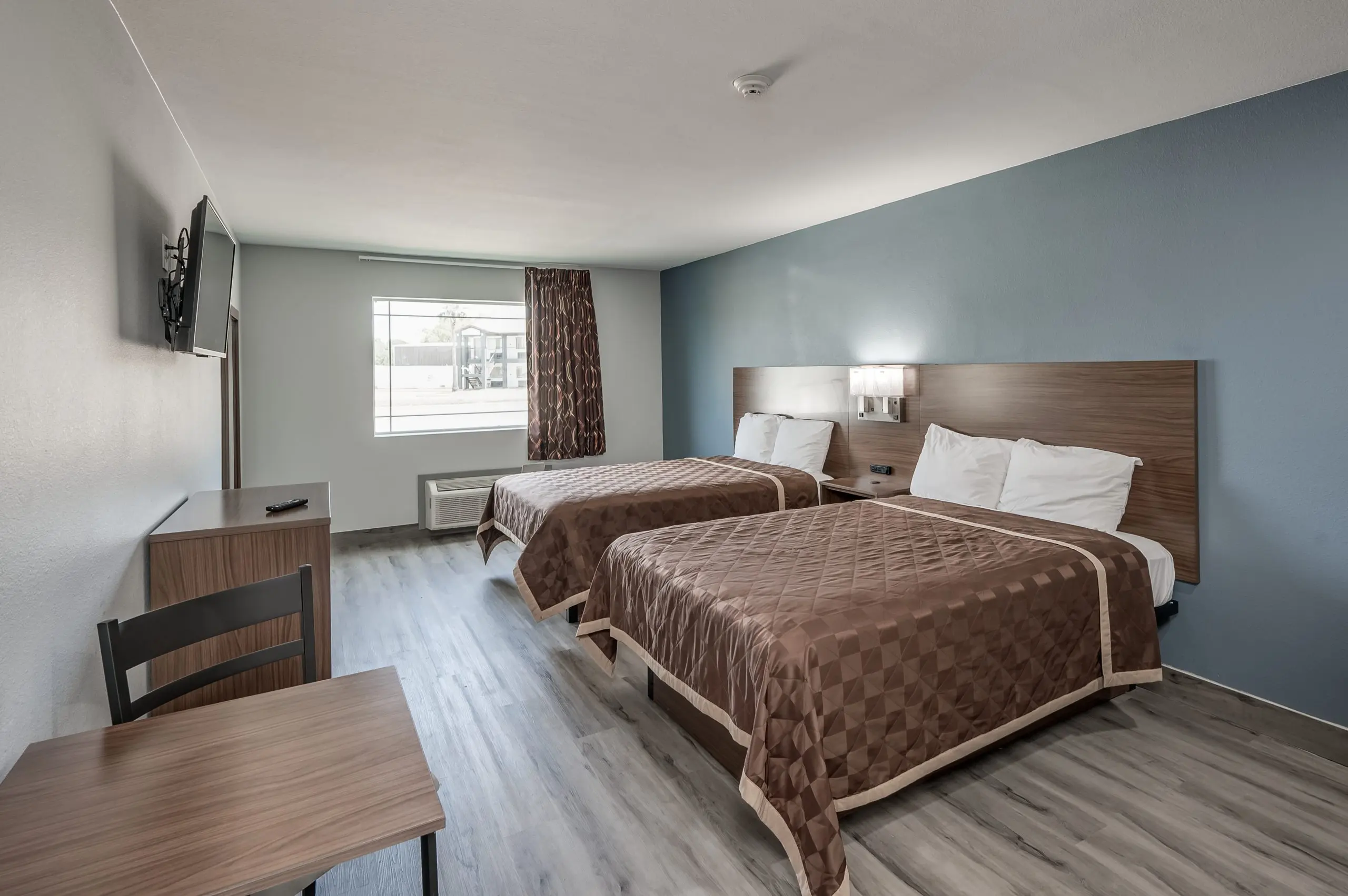 A spacious hotel room featuring two beds, a desk, and a television mounted on the wall. The room has a large window allowing natural light, with one side decorated in light blue and the other in neutral colors.