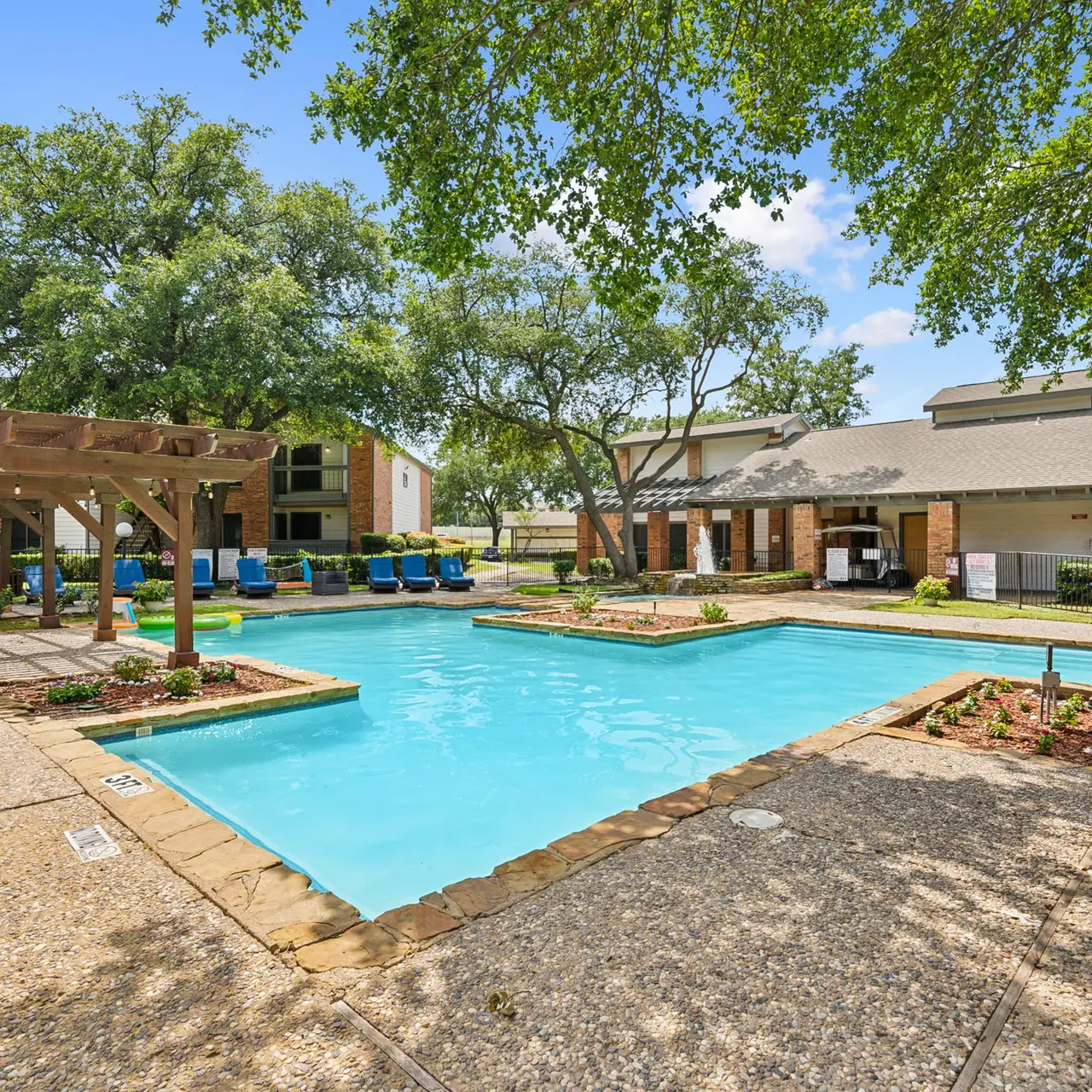 A serene pool area surrounded by trees and residential buildings, featuring a clear blue pool, lounge chairs, and shaded picnic tables.