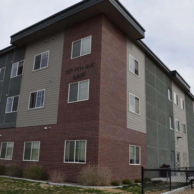 Modern Apartment Building A modern three-story apartment building with a combination of brick and green siding. The building has multiple windows and is set against a cloudy sky with some greenery visible in front.