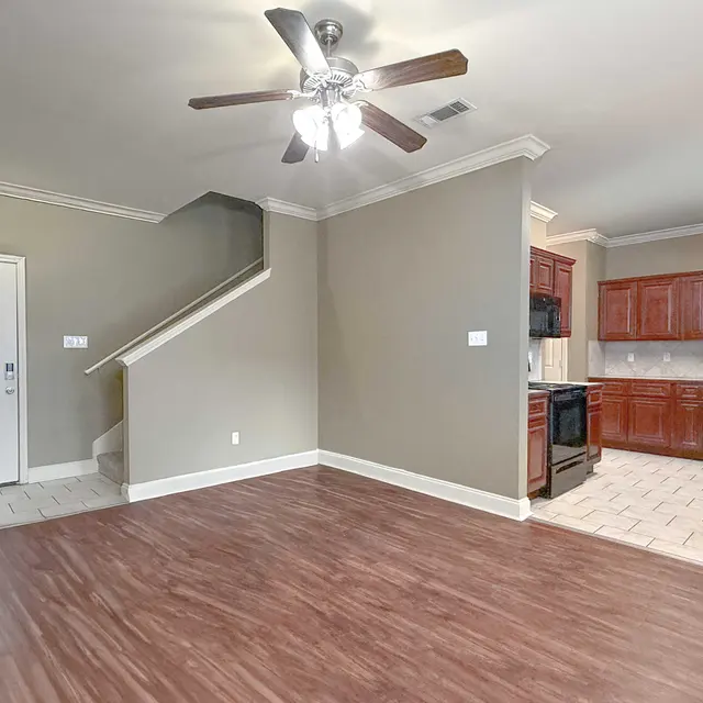 A spacious living room area featuring a ceiling fan, hardwood flooring, and a kitchen area with wooden cabinetry. A staircase leads to an upper level. The room has neutral wall colors and a door leading outside.