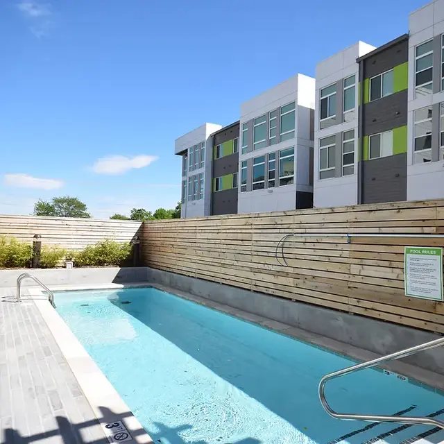 A modern pool area featuring a rectangular swimming pool, surrounded by wooden fencing, with lounge chairs on one side and residential buildings in the background.