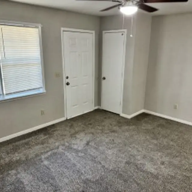 An empty room featuring beige walls and a ceiling fan. A large window with blinds allows natural light in, and there are two doors: one leading outside and one possibly leading to a closet.