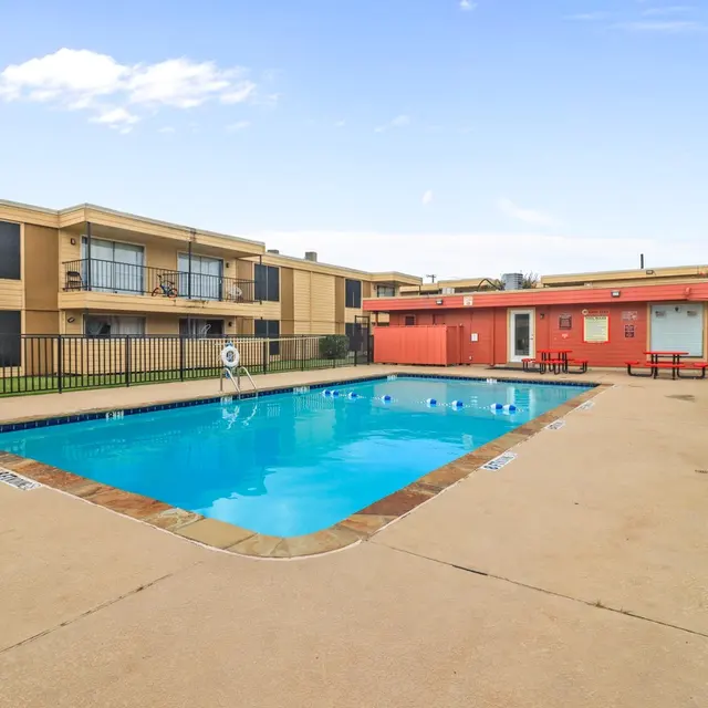A clear swimming pool surrounded by outdoor concrete space and apartment buildings. Bright blue sky above.