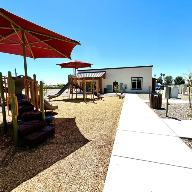 Sunny Playground with Shade Structures A sunny playground featuring a play structure with slides, surrounded by gravel, with two large shade umbrellas nearby. In the background, a building is visible.