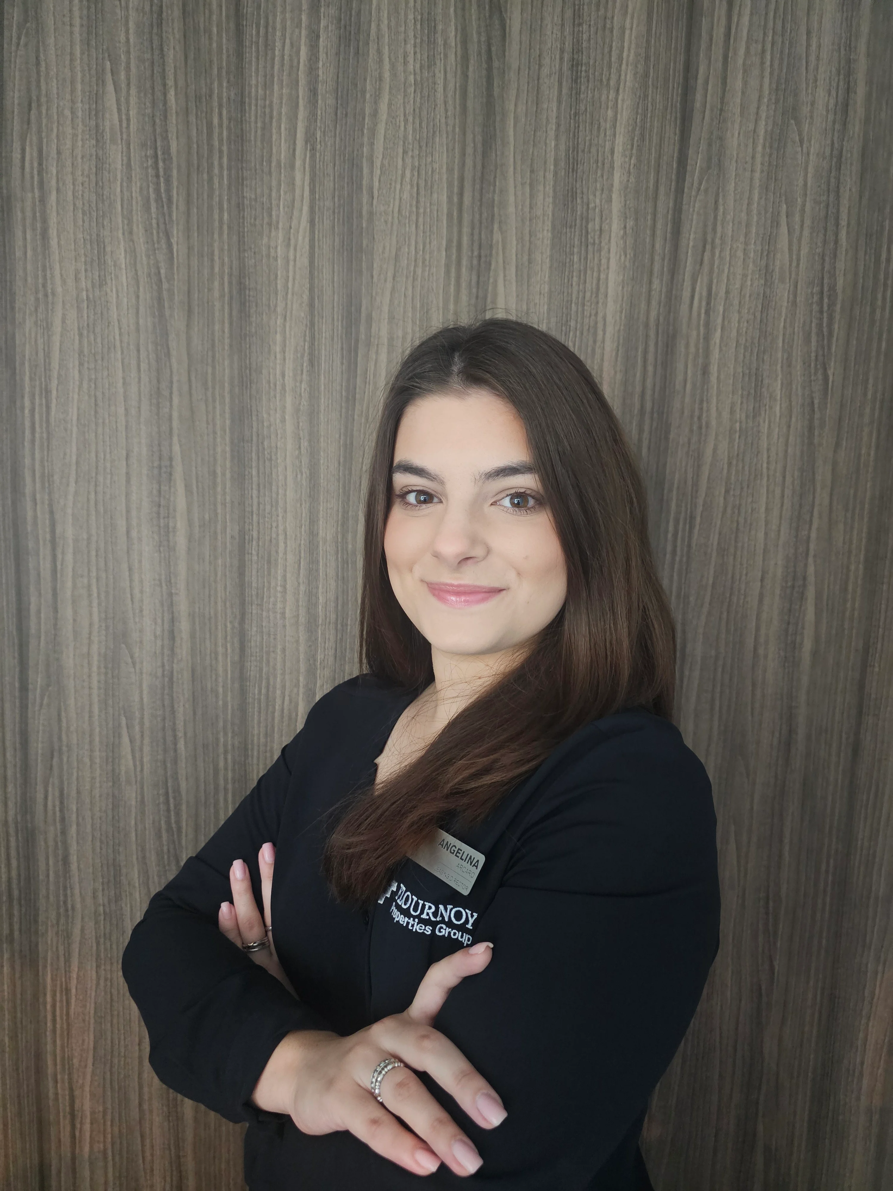Confident Businesswoman A woman with long brown hair, wearing a black blazer, stands confidently with her arms crossed in front of a wooden background.