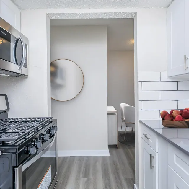 A modern kitchen with stainless steel appliances, including a gas stove and microwave, white cabinets, and a gray countertop. A bowl of red apples is on the countertop, and an adjacent room is partially visible through an open doorway.