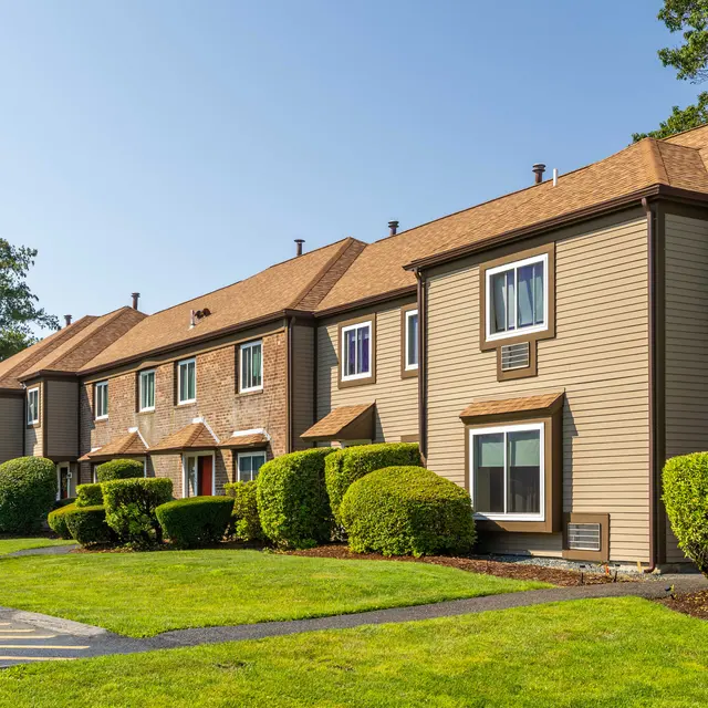 A row of townhouses with flat roofs and beige siding. Each unit has large windows and a small front yard with trimmed bushes. A clear blue sky is in the background, suggesting a sunny day.