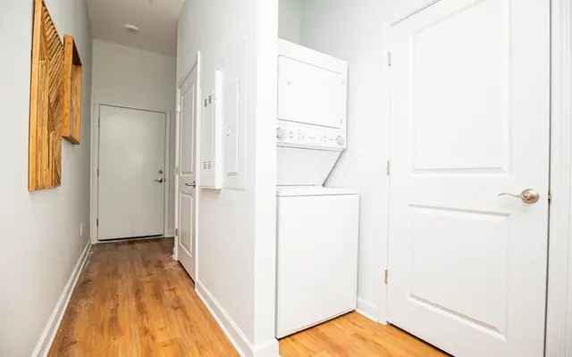 A bright hallway featuring white walls and wooden flooring, with a washing machine and dryer unit located on one side. A decorative wooden piece is mounted on the wall, and a door is visible at the end of the hallway.