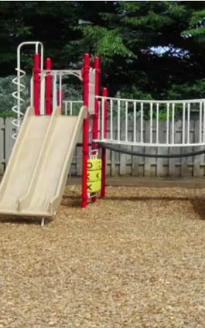 A playground featuring two slides, one beige and one red, surrounded by wood chip ground cover and a wooden fence in the background.