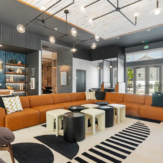 A modern lounge area featuring a large orange sectional sofa, two white and one black coffee tables on a patterned rug, and elegant lighting fixtures above. The space includes a bar area with a blue accent wall and decorative shelves, with large windows allowing natural light to enter.