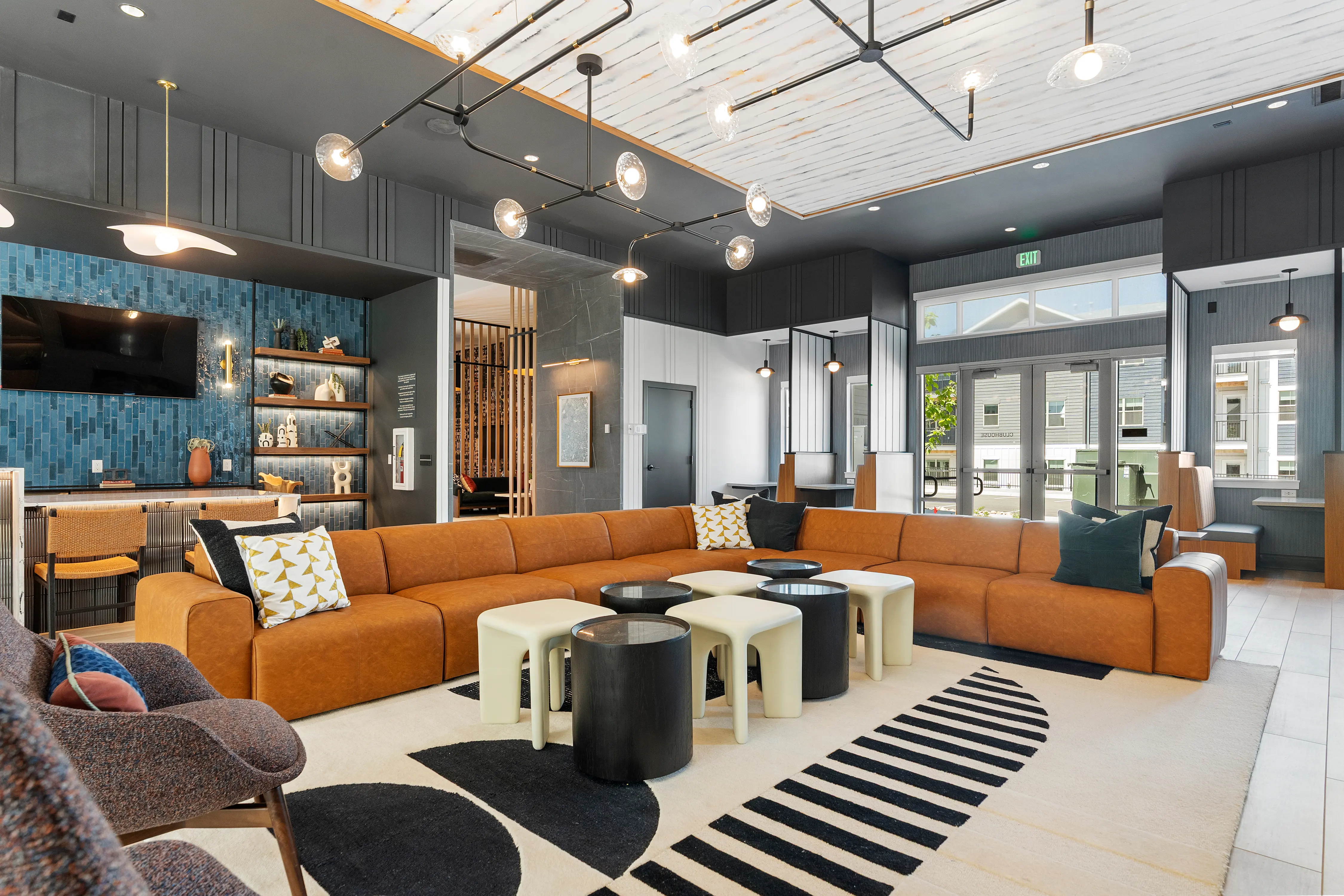 A modern lounge area featuring a large orange sectional sofa, two white and one black coffee tables on a patterned rug, and elegant lighting fixtures above. The space includes a bar area with a blue accent wall and decorative shelves, with large windows allowing natural light to enter.