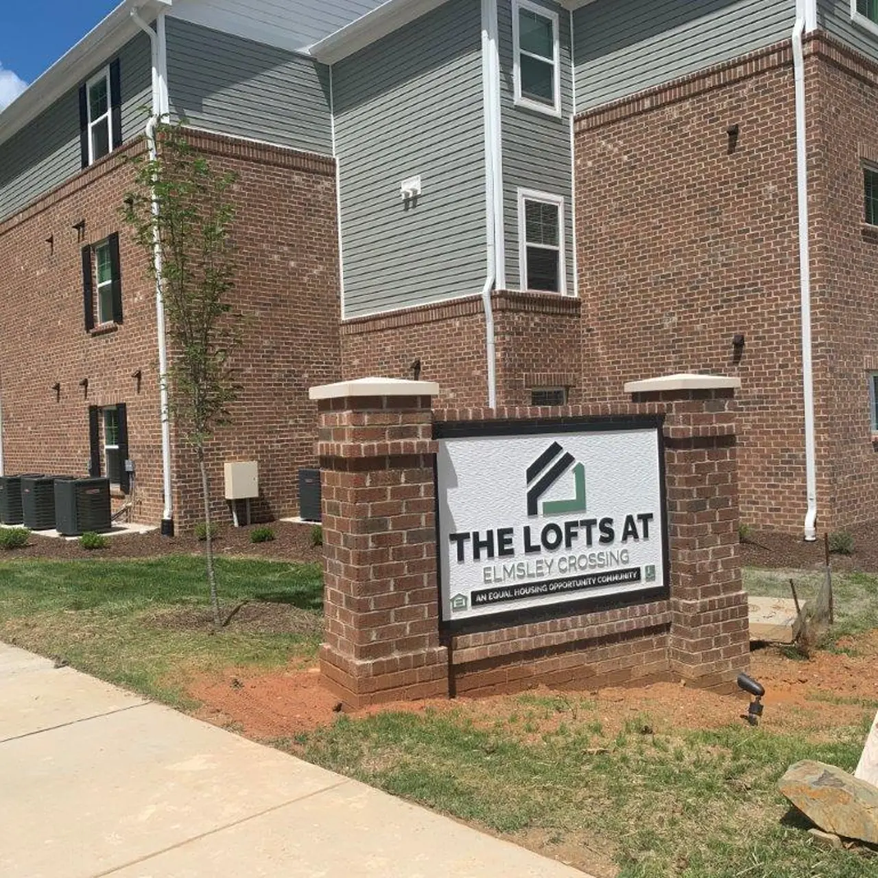 Sign marking the entrance to 'The Lofts at Elmley Crossing,' with a brick feature and landscaping in front of a residential building.