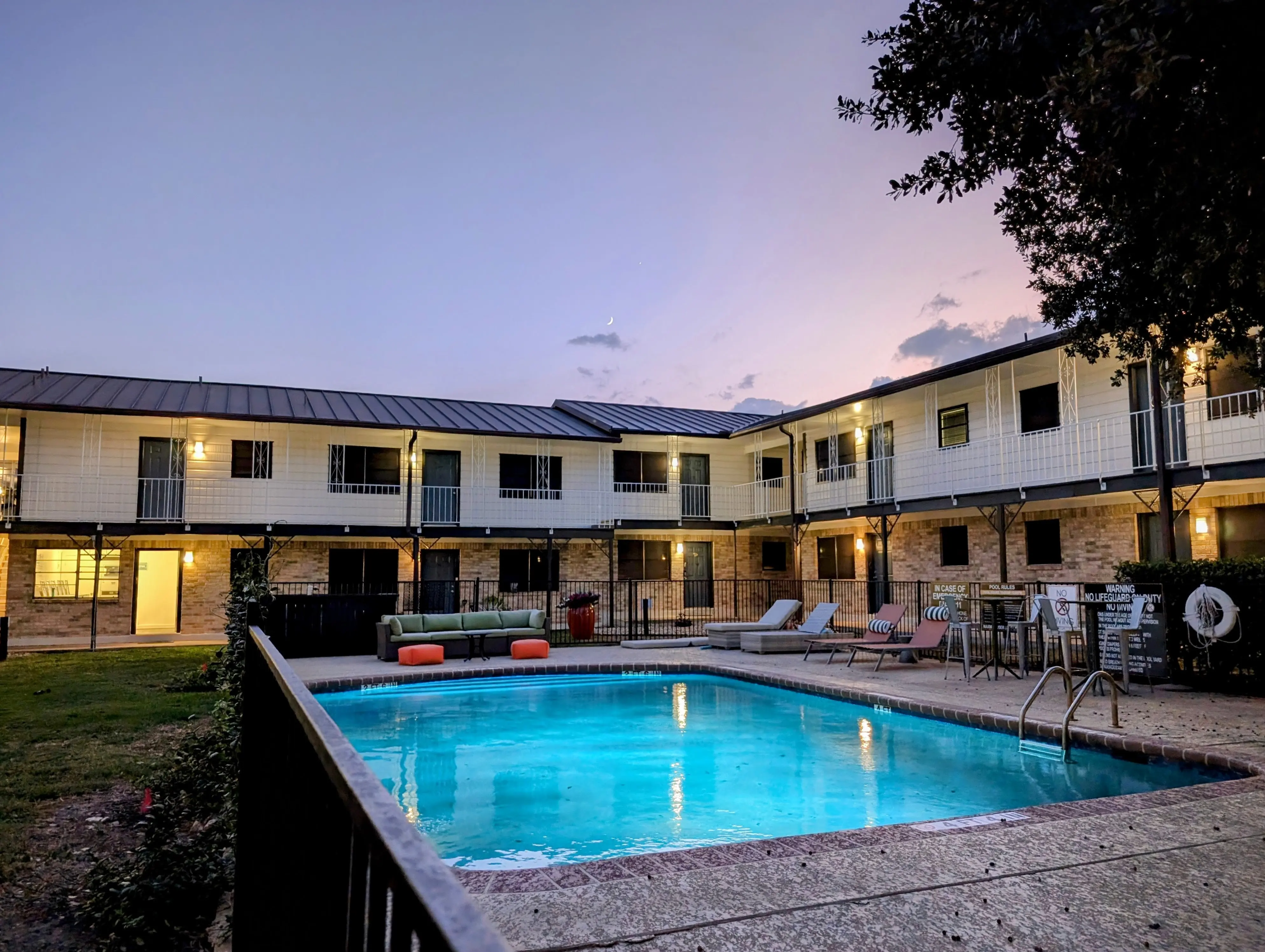 An outdoor swimming pool surrounded by lounge chairs and a grassy area, with a two-story apartment building in the background during twilight.