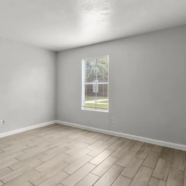 A vacant room with light grey walls and wooden tile flooring. There is a window on one wall allowing natural light to enter.