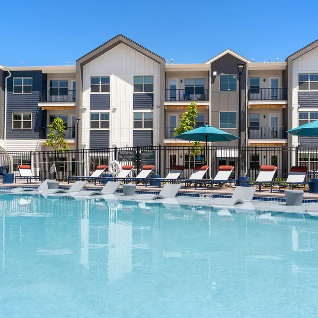 A modern apartment complex with a swimming pool. Lounge chairs lined up near the pool, with umbrellas providing shade. The building features a mix of textures and colors, with balconies visible.