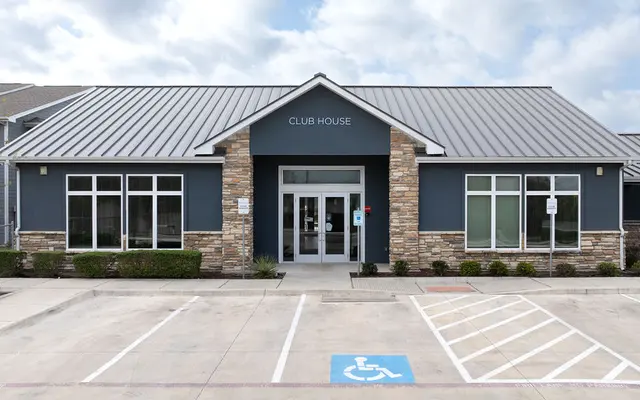Exterior view of a modern clubhouse, featuring a stone and blue exterior with large windows and a metal roof, set against a cloudy sky.