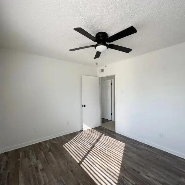 Bright Empty Room A bright, empty room with a ceiling fan and hardwood flooring. The walls are white and there's a window with blinds allowing sunlight to stream in, casting shadows on the floor. A doorway is visible in the background leading to another part of the space.