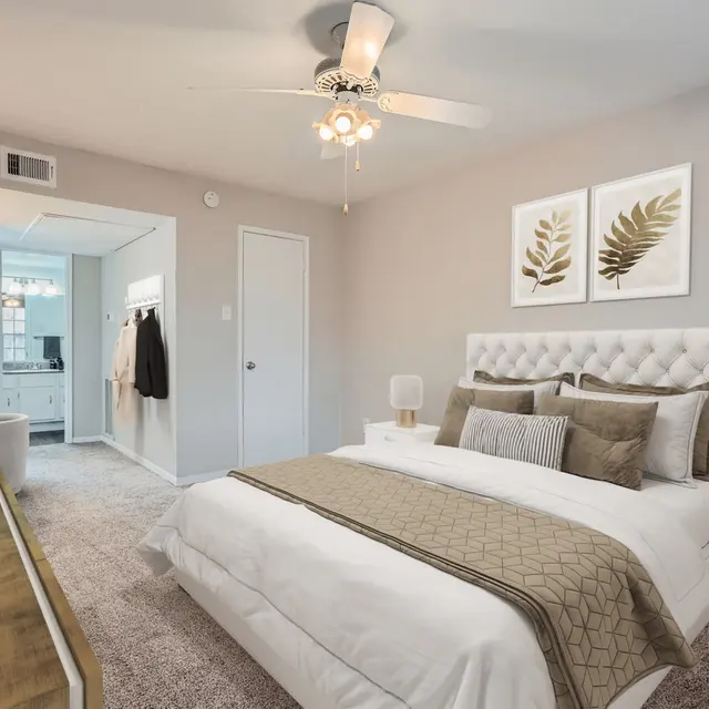 A modern bedroom with a queen-sized bed dressed in white and beige linens, featuring a tufted headboard. There are decorative pillows on the bed, a nightstand on each side with lamps, and a neutral-toned decor. A doorway leads to a bathroom area visible in the background, and the walls are painted in a soft gray color. There are framed botanical prints above the headboard.
