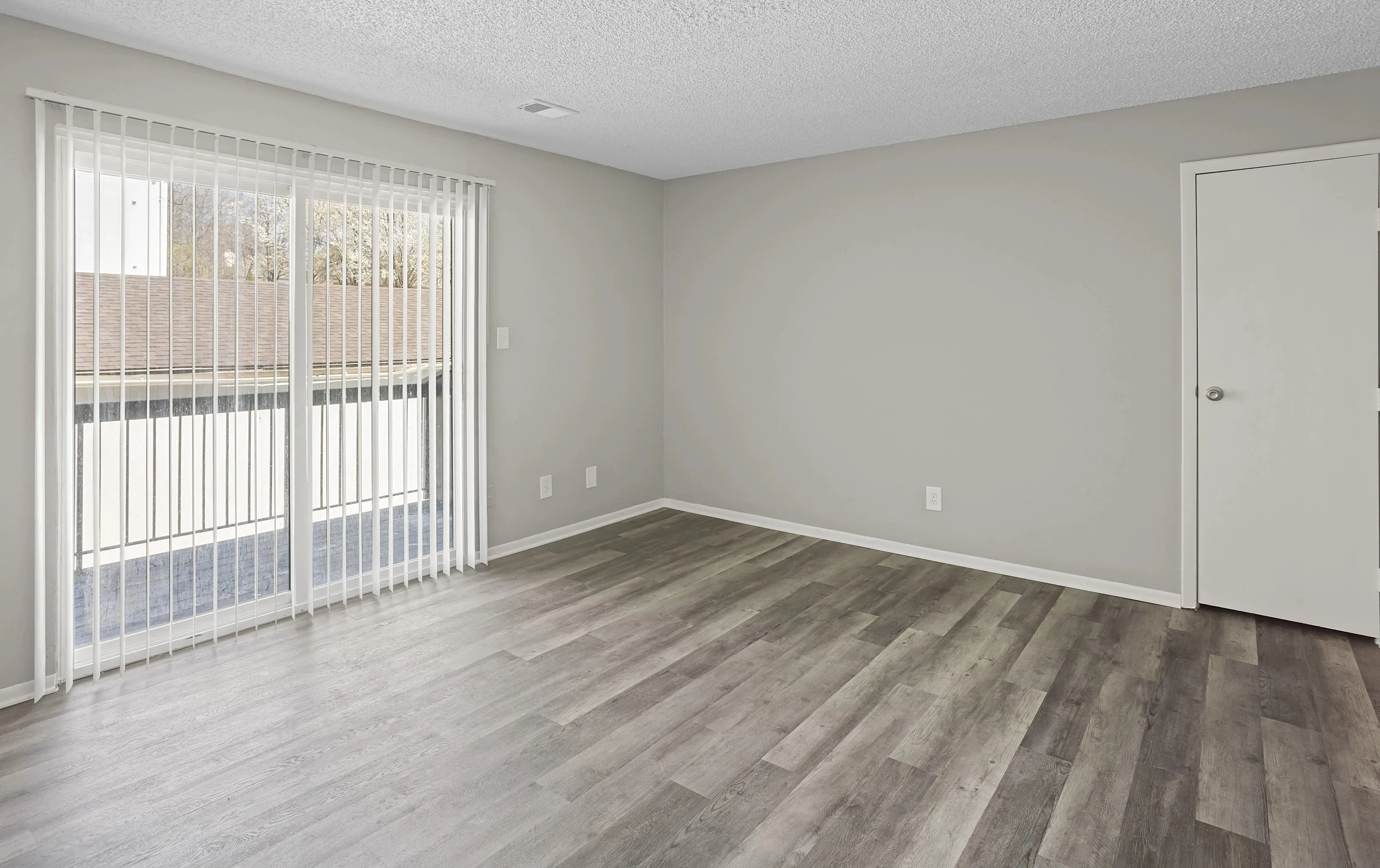 An empty living room featuring light wood flooring, beige walls, and large sliding glass doors with vertical blinds. A single door is visible on the right side of the room.