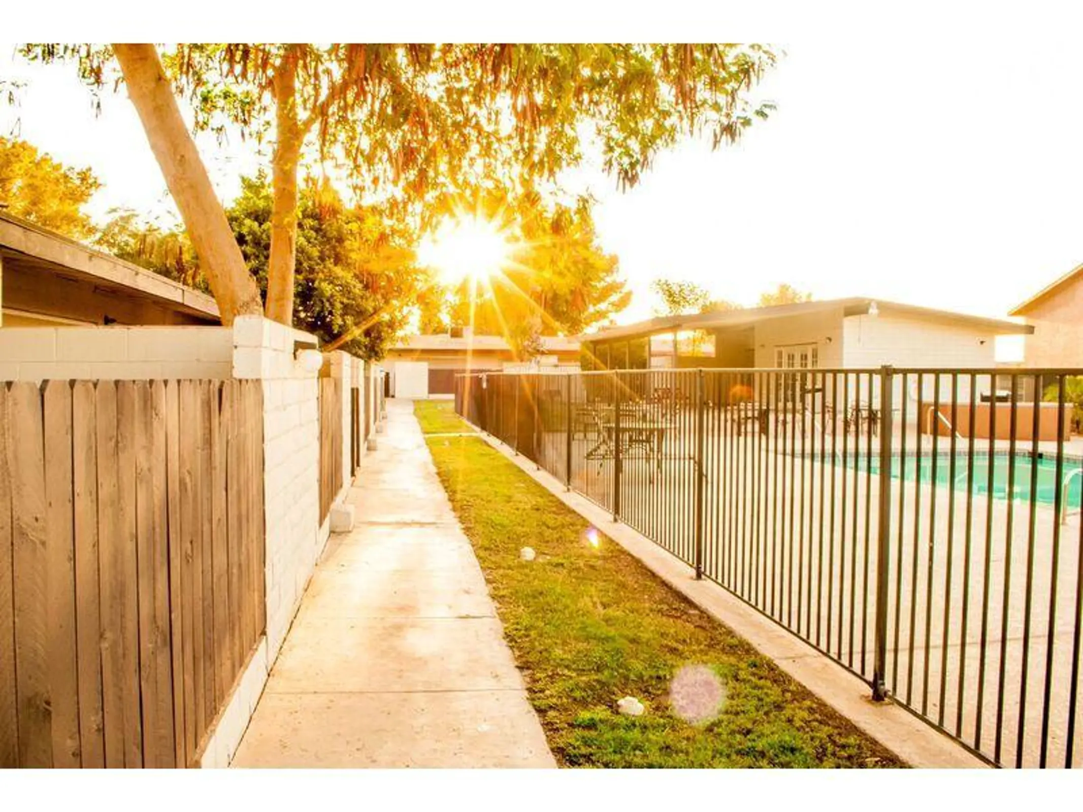 Sunlit walkway beside a fenced swimming pool and apartment buildings.