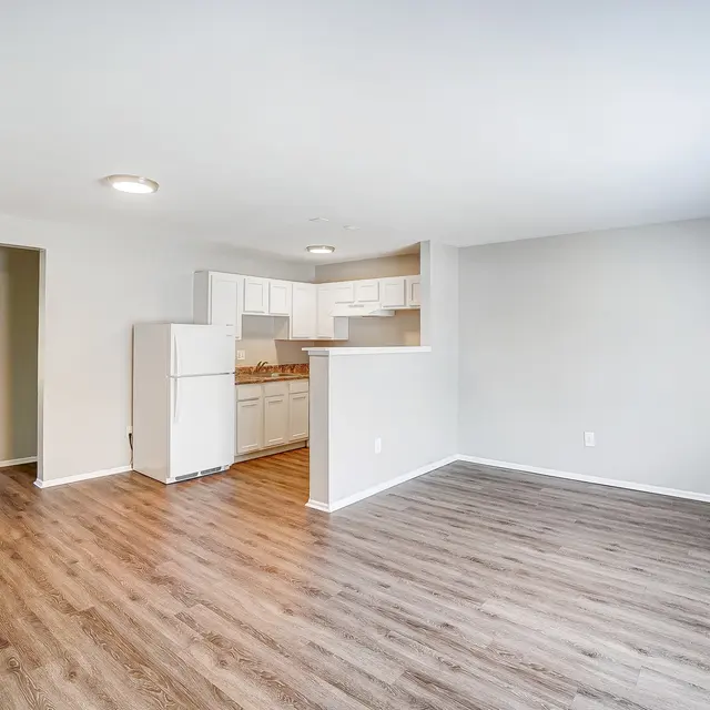 A spacious empty apartment featuring wooden flooring, an open layout connecting the living area and kitchen, and a doorway leading to another room.
