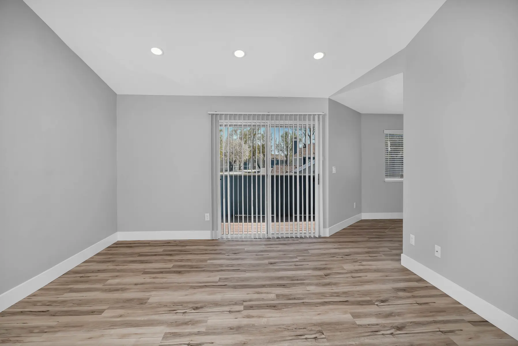 A modern living room with gray walls and wooden flooring, featuring a sliding glass door with vertical blinds that leads to an outside area. The room has recessed lighting and a corner window allowing natural light.