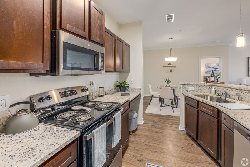 A modern kitchen with dark wooden cabinets, stainless steel appliances, and granite countertops. The kitchen features a stove, microwave, sink, and a dining area in the background.