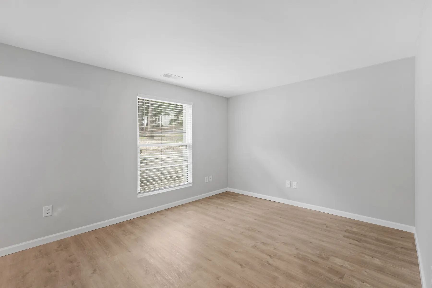 A vacant room with light gray walls and wooden flooring. A window with white blinds lets in natural light, illuminating the space.