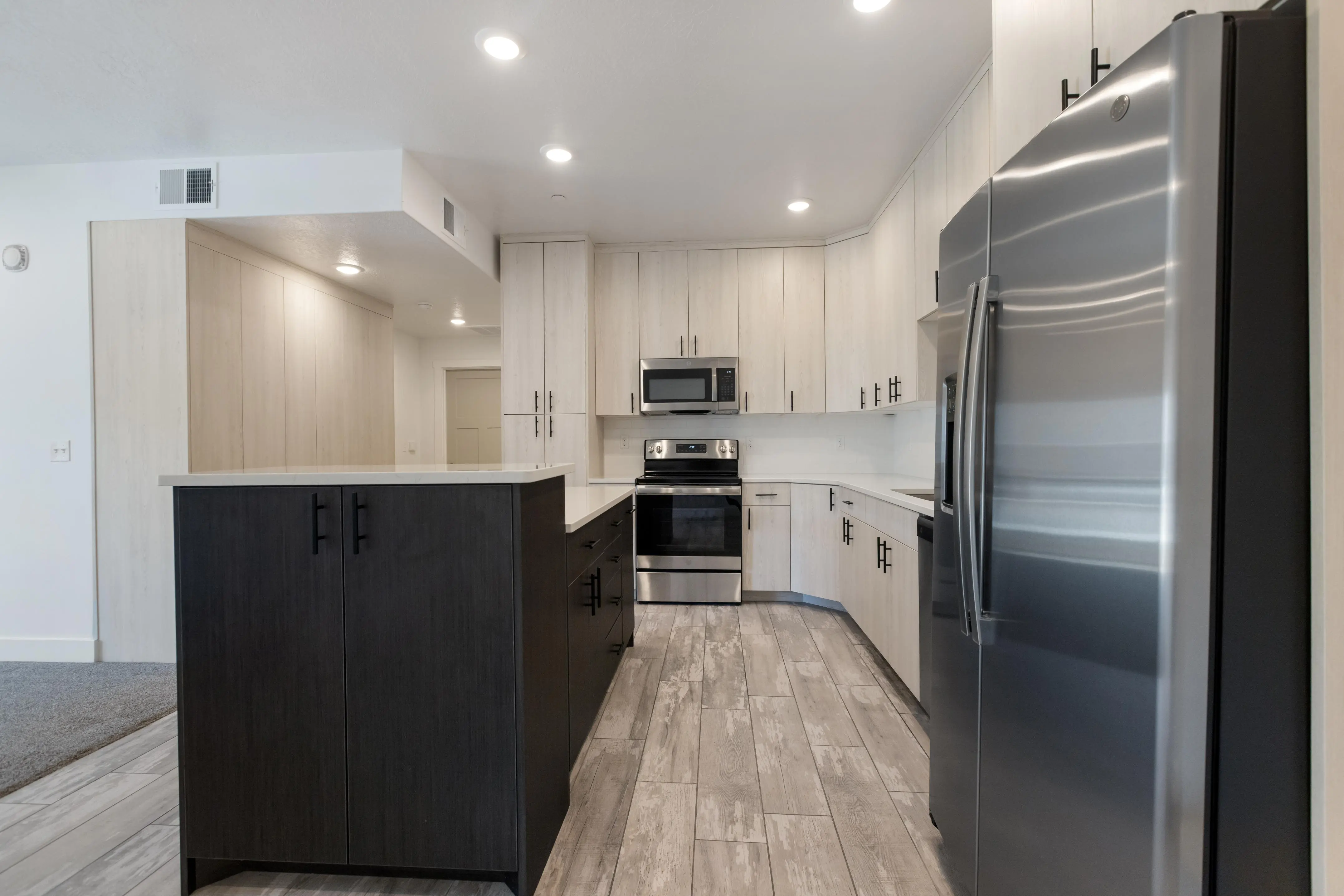 A spacious modern kitchen featuring a dark kitchen island, stainless steel appliances, and light-colored cabinets with a combination of neutral tones in the flooring.