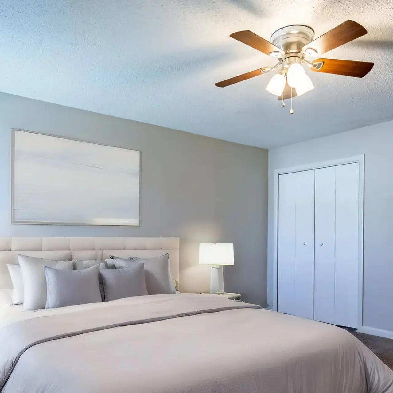 A cozy modern bedroom featuring a large bed with light gray bedding, two bedside lamps, a TV on a white dresser, and a ceiling fan with wooden blades. The walls are painted light blue, and there is artwork on the wall. A white chair is positioned near the bed, and there are double doors leading to another room.