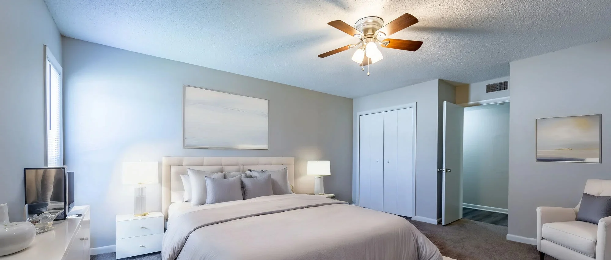 Modern Bedroom Interior A cozy modern bedroom featuring a large bed with light gray bedding, two bedside lamps, a TV on a white dresser, and a ceiling fan with wooden blades. The walls are painted light blue, and there is artwork on the wall. A white chair is positioned near the bed, and there are double doors leading to another room.