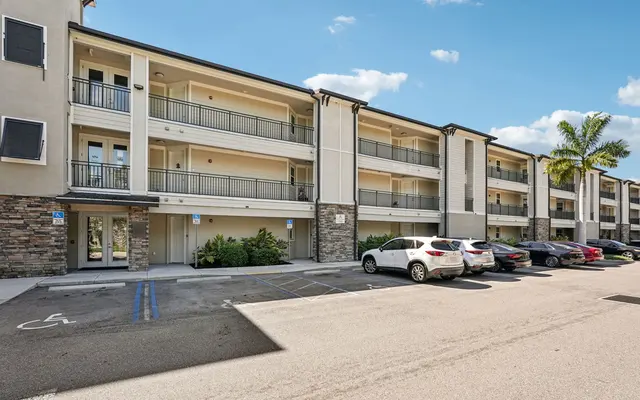 Modern Apartment Building A modern three-story apartment building with multiple balconies and a parking lot in front. Several vehicles are parked, and there are palm trees nearby.