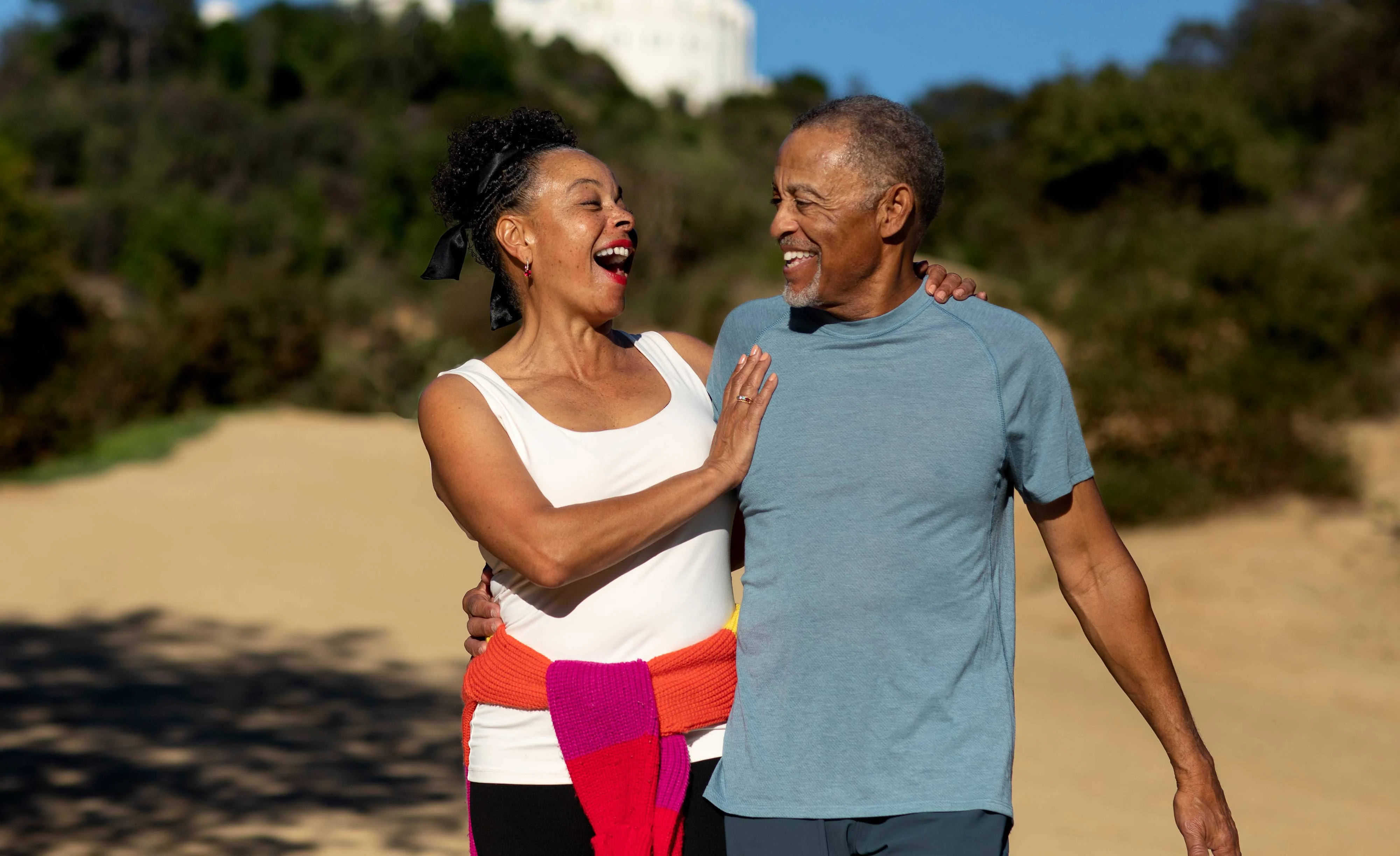 Joyful Couple Walking Outdoors A joyful couple walking together in a sunny outdoor setting. The woman, wearing a white tank top and black leggings, is laughing and playfully touching the man's shoulder. The man, in a blue shirt and light grey shorts, smiles back at her. They are on a dirt path with greenery in the background.