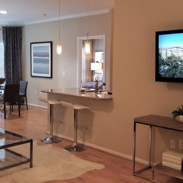 A modern apartment interior featuring a living area with a black leather couch, a coffee table, and a television mounted on the wall. There is a dining area with a round table and chairs in the background and large windows with sheer curtains. The decor is minimalist with a light-colored rug and wooden flooring.
