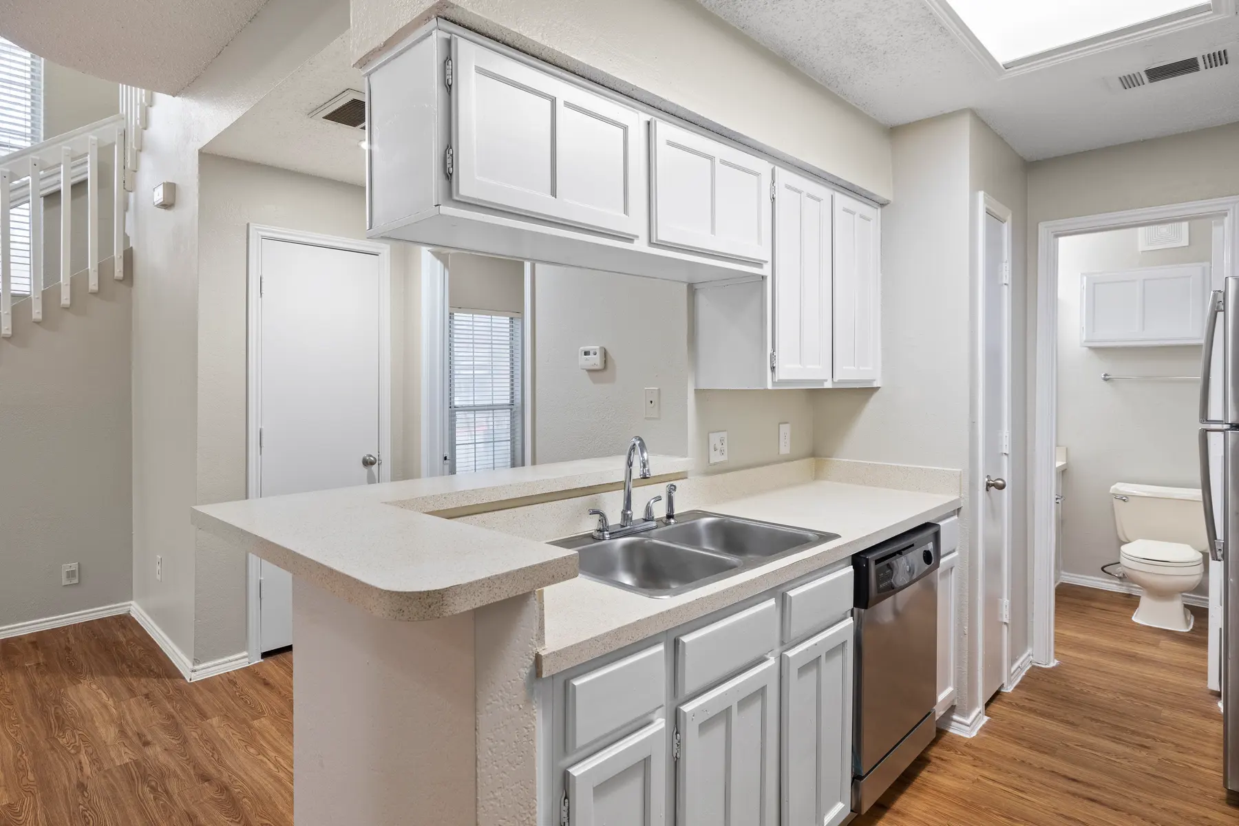 A modern kitchen with white cabinetry and stainless steel appliances. The kitchen features a double sink, dishwasher, and an open layout that connects to the living area. There is a hallway leading to a bathroom and a staircase in the background.