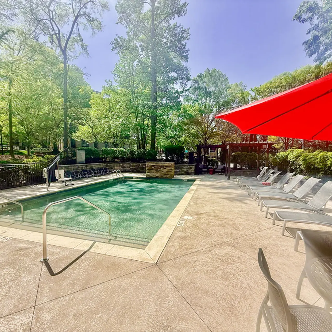 A serene swimming pool area surrounded by trees with lounge chairs and a bright red umbrella.