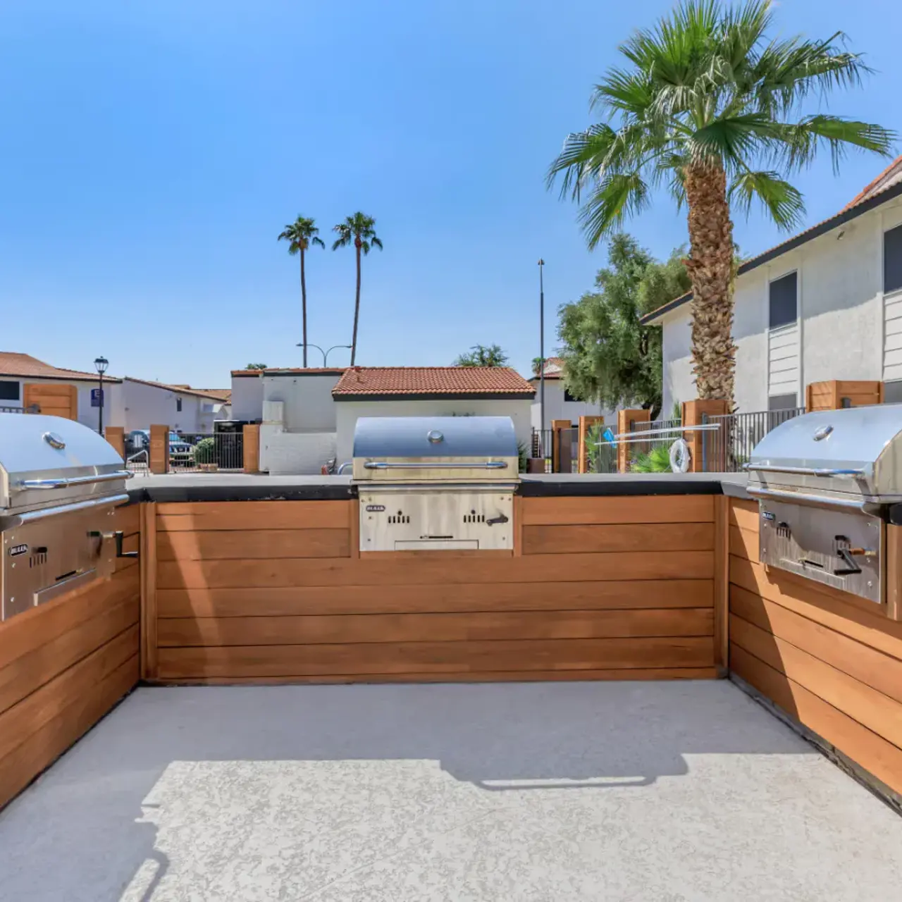 A modern outdoor grilling area featuring stainless steel grills and wooden siding, with palm trees in the background and clear blue skies.
