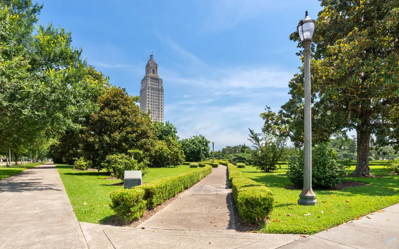 A landscaped park pathway leading towards a tall building in the background under a clear blue sky. The scene features well-maintained grass, trees, and bushes along the walkway.