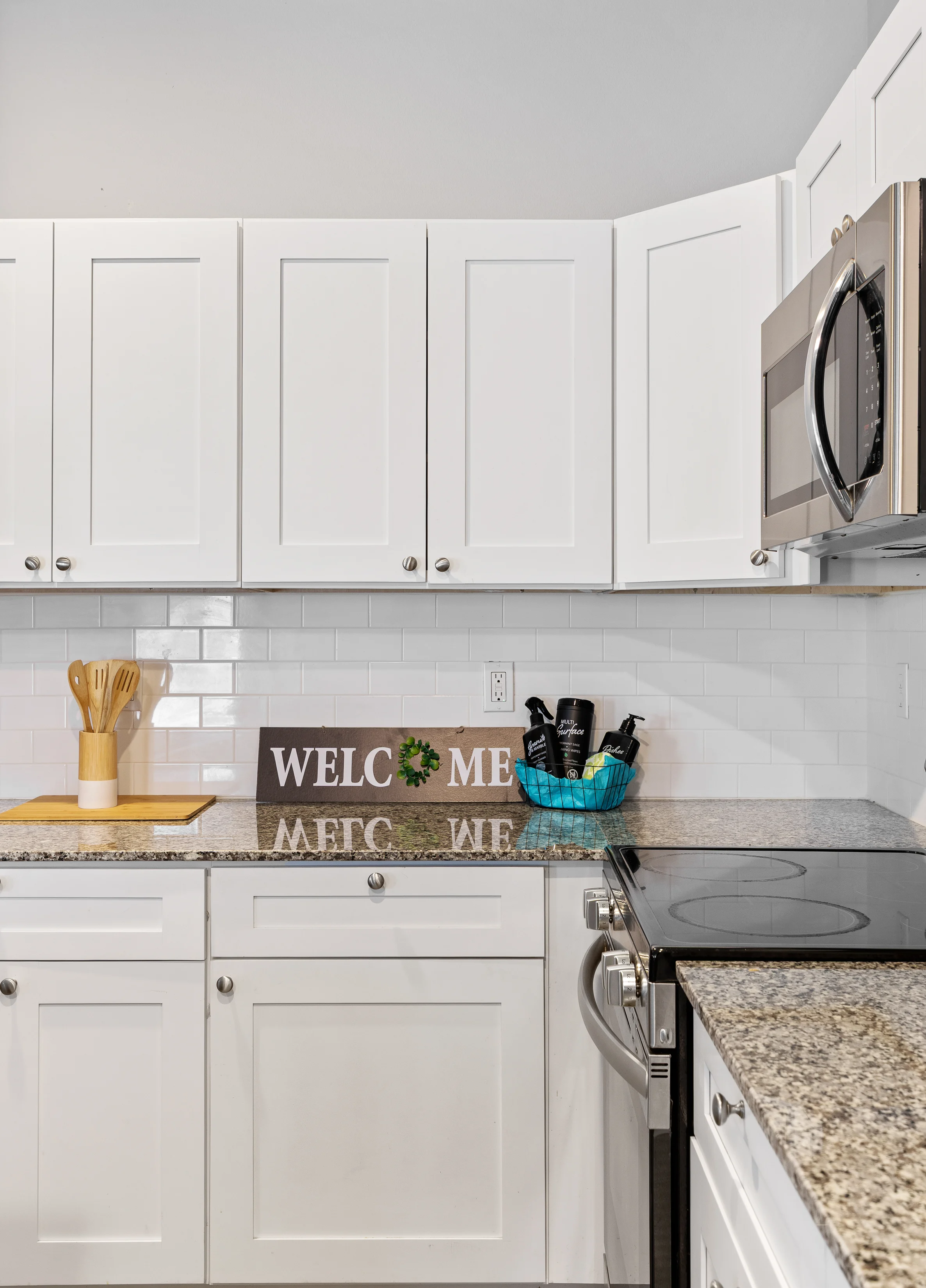 Stylish Modern Kitchen A modern kitchen featuring white cabinets, a granite countertop, a stainless steel microwave, and a decorative sign that reads 'WELCOME.' There are also kitchen utensils and a utensil holder on the countertop.