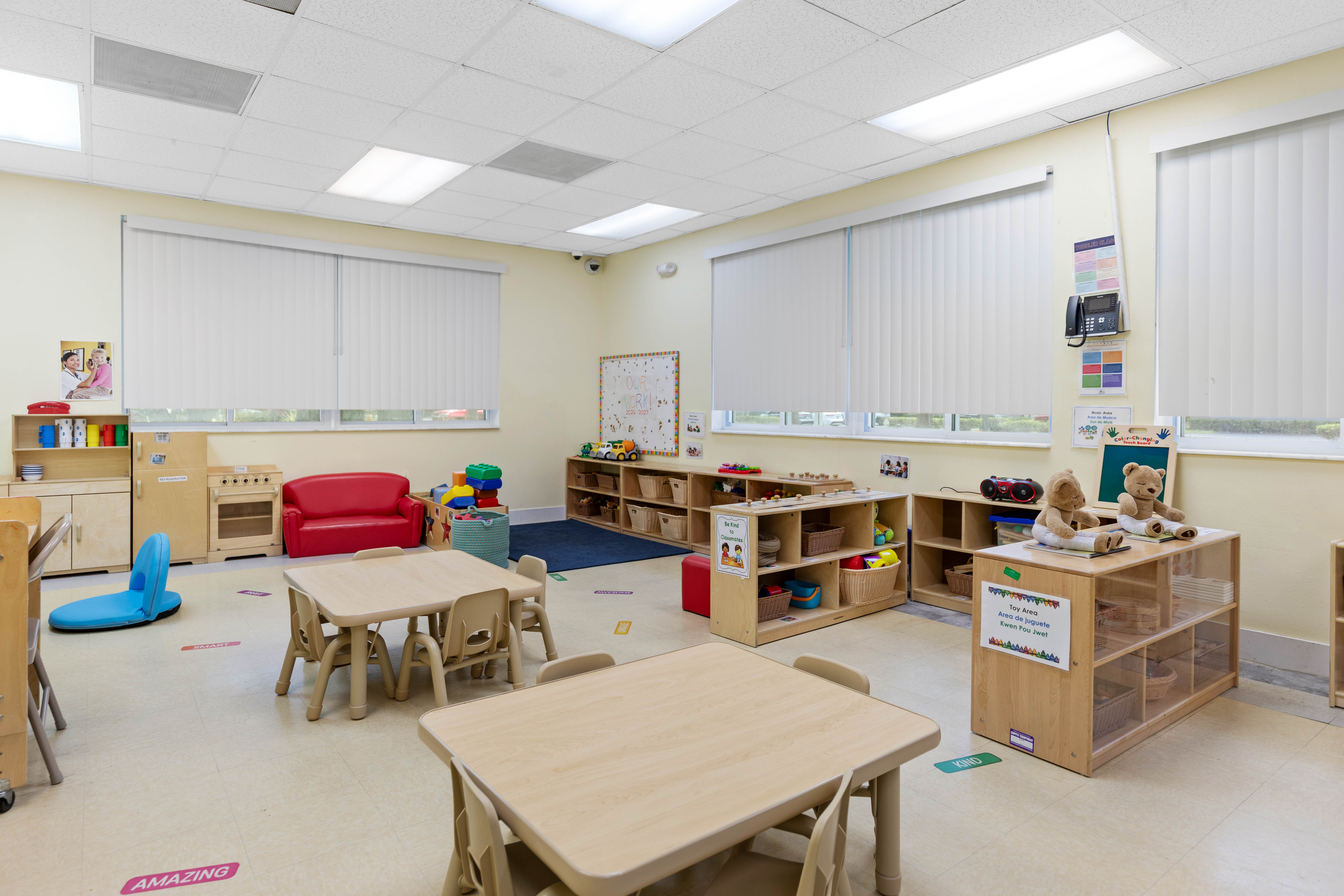 Bright Classroom Interior A bright and tidy classroom featuring wooden tables and chairs, a red chair, and shelves filled with toys and learning materials. Large windows allow natural light to fill the space.