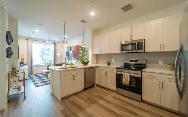 A modern kitchen with wooden flooring, featuring light-colored cabinets, stainless steel appliances, and a kitchen island with seating. Large windows allow natural light to fill the space, complemented by decorative plants and contemporary lighting fixtures.