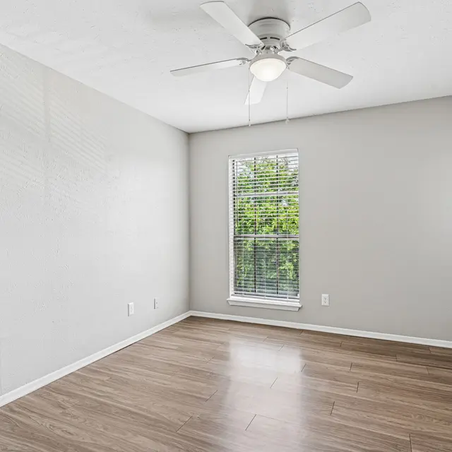 An empty room featuring wooden flooring, a ceiling fan, and two windows with blinds allowing natural light to enter.