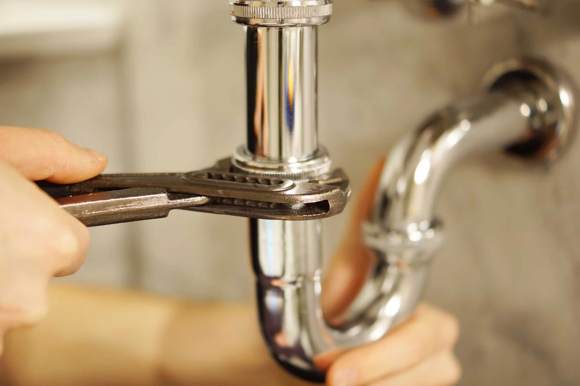 A person using an adjustable wrench to tighten a shiny chrome pipe under a sink