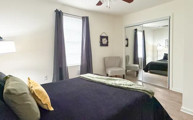 A cozy bedroom featuring a black bedspread with decorative pillows, a nightstand with a lamp, and dark curtains. In the reflection, another room with a green bed can be seen, along with a chair by a window.
