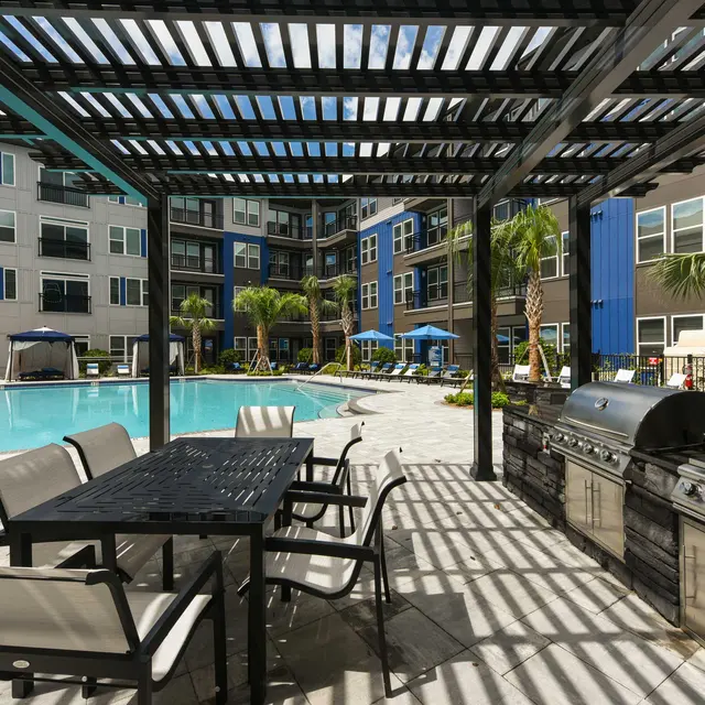 Elegant Apartment Pool Area with BBQ A spacious outdoor area featuring a swimming pool surrounded by palm trees, lounge chairs, and a modern grill under a pergola.