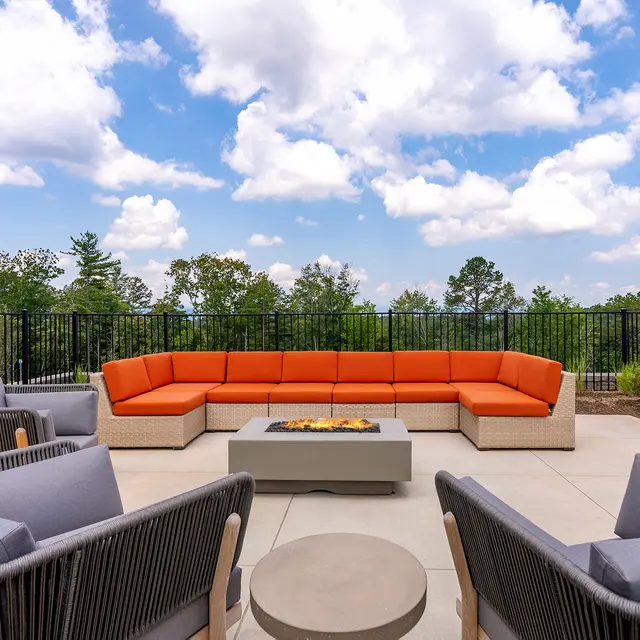 A modern outdoor lounge area featuring an orange sectional sofa, grey armchairs, and a fire pit on a patio with green trees in the background.